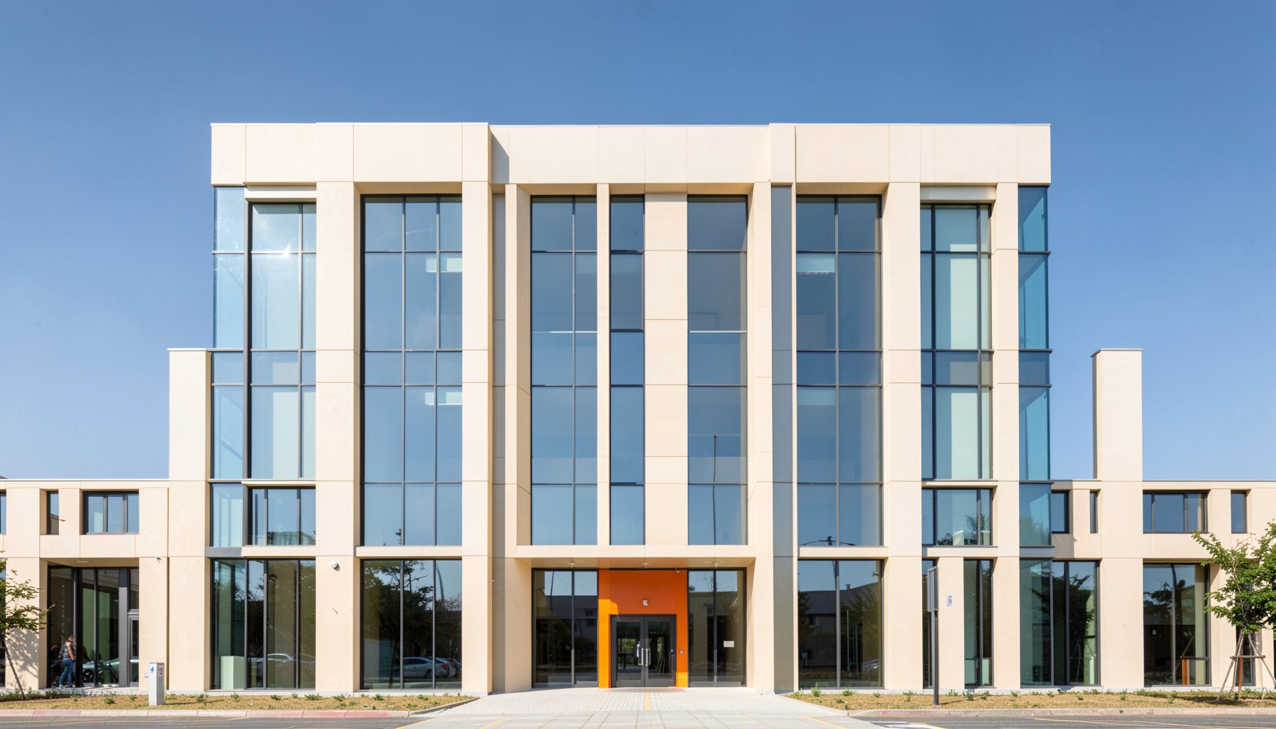 صورة معمارية نظيفة لمبنى أكاديمي حديث بتصميم متماثل، واجهة باللون البيج الفاتح مع نوافذ زجاجية عاكسة، سماء زرقاء صافية في الخلفية